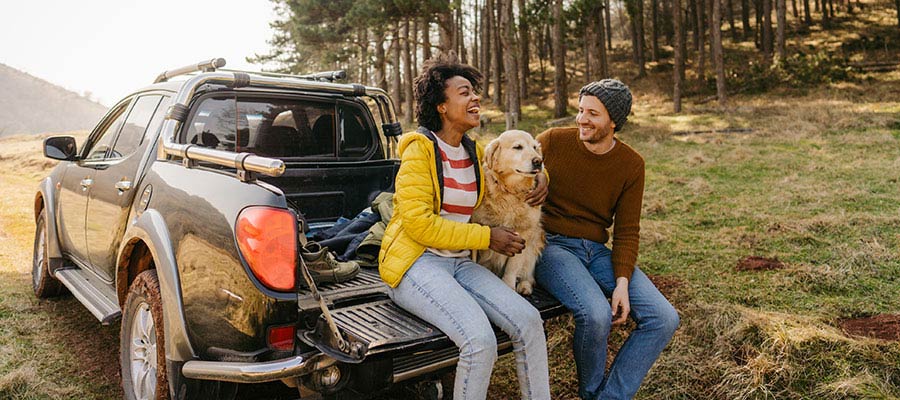 Couple outdoor sitting on back of parked truck hugging their golden retriever