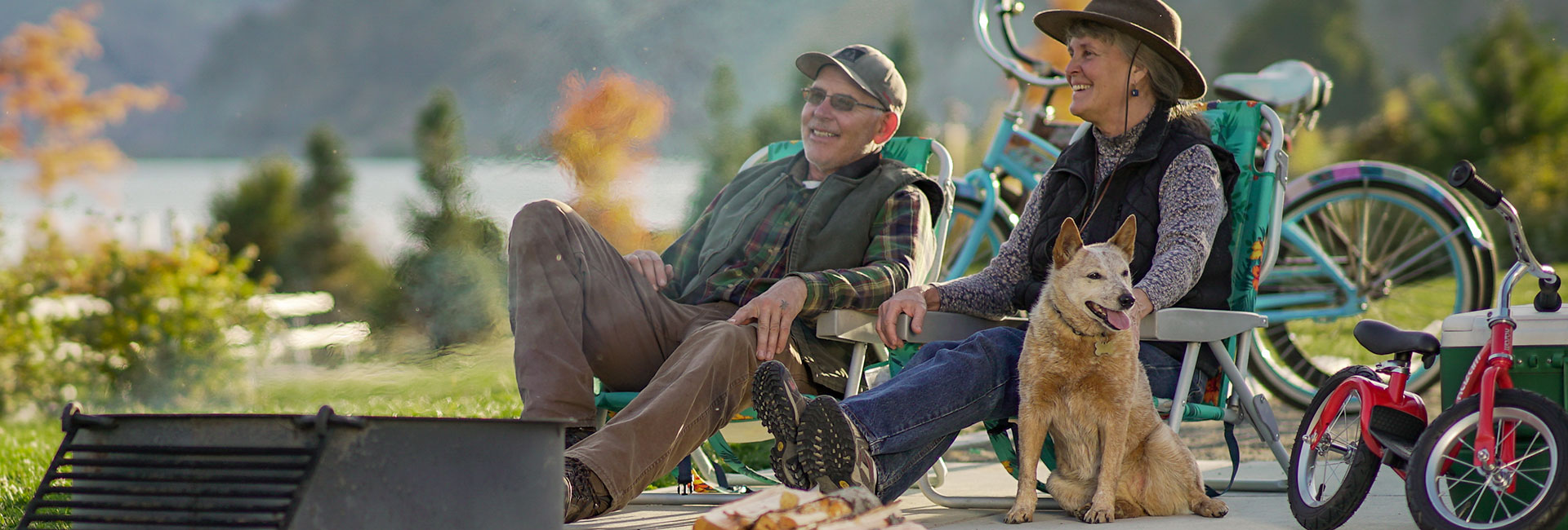 A man and woman with their dog sitting by an outdoor fire next to a beautiful lake in the PNW